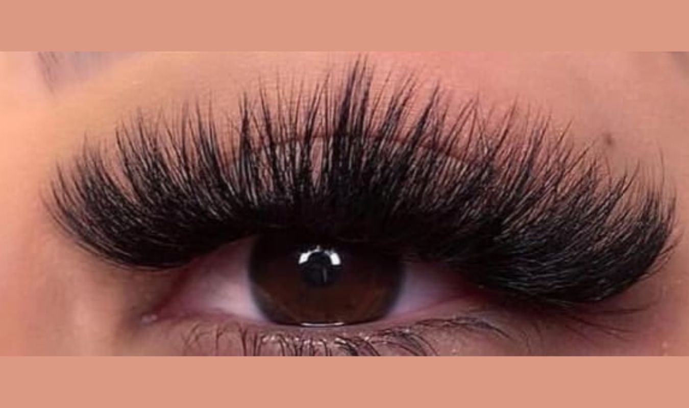 Mega Volume Lash Extension Transform your look with Stunning lashes 1 Close-up of a brown eye with long, thick false eyelashes, surrounded by skin. A few natural eyelashes are visible beneath the false ones, with a soft, muted orange background.