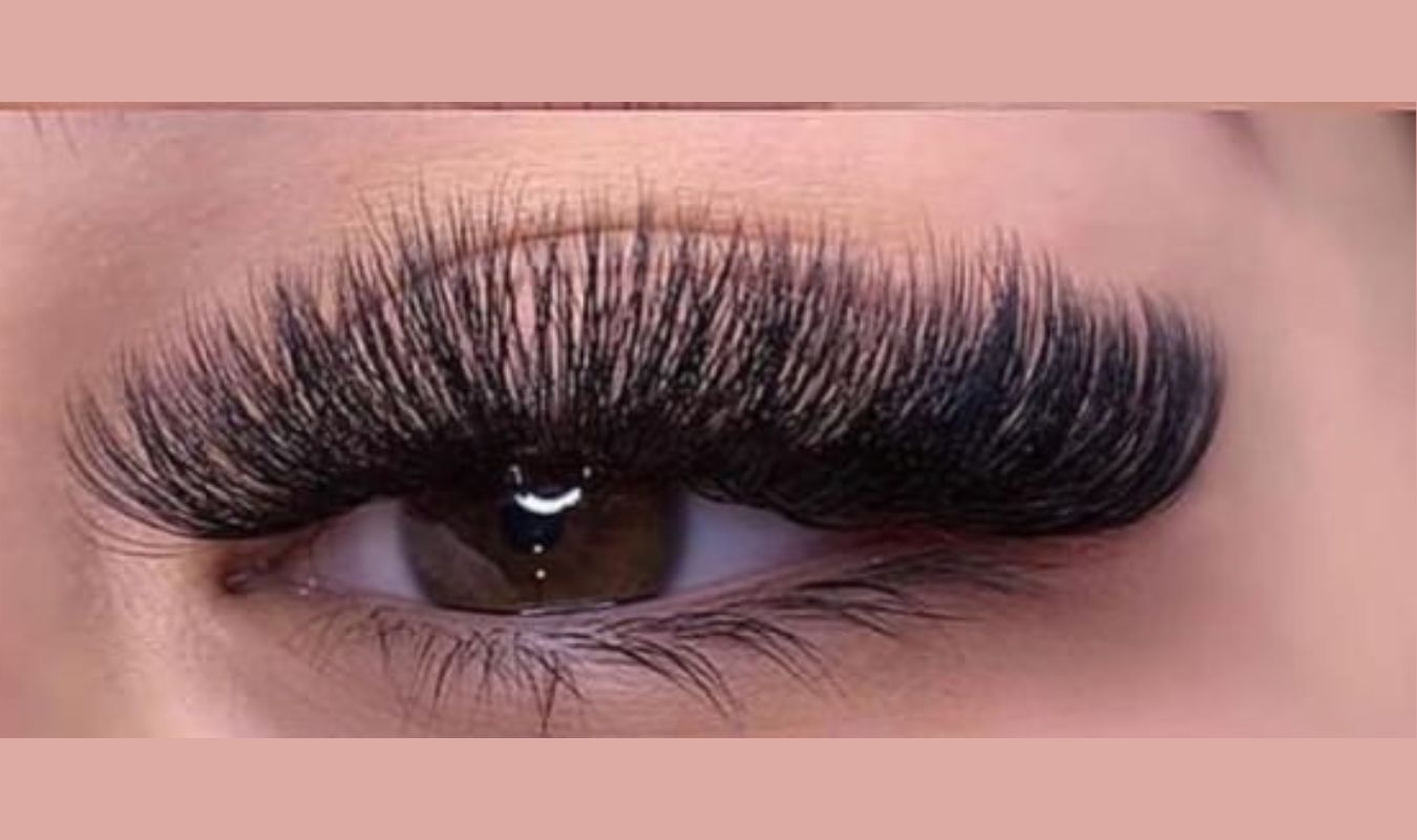 Close-up image of an eye with dramatic, long, black false eyelashes. The eye is brown, and the eyelid is smooth and well-defined, showing a subtle sheen. The background is a soft, muted pink, enhancing the focus on the eye and lashes.