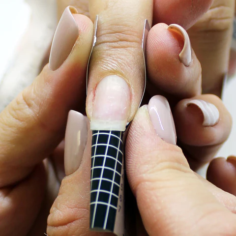 A close-up of a hand receiving a gel nail extension. A technician is applying a nail form to the natural nail to shape and support the gel application. The natural nail appears unpolished and is situated between the technician's fingers. Surrounding nails are painted in soft, neutral tones