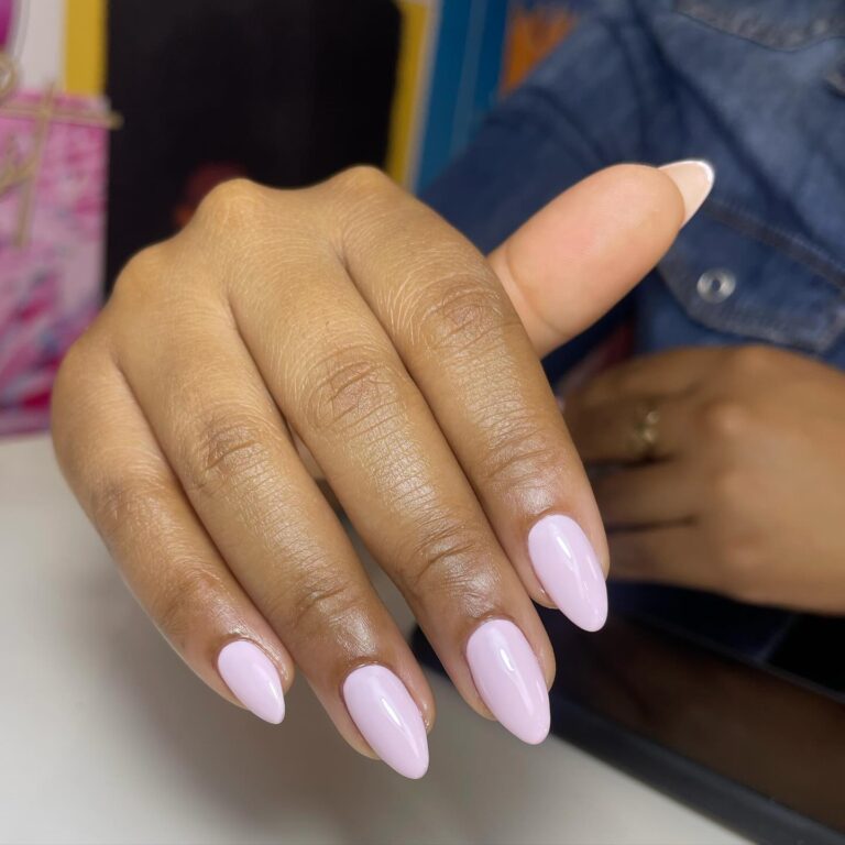 Nail Salons Nairobi: Get the Best Manicure & Pedicure Services. 7 A close-up of a hand displaying long, almond-shaped nails painted in a soft pastel pink color. The hand is resting on a flat surface, with a slender finger pointing upwards. The background includes blurred colorful images and a denim jacket sleeve is visible on the right side of the image.