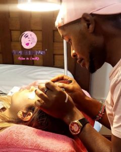 A beauty professional, wearing a pink cap and shirt, gently applies eyelash extensions to a client lying on a treatment bed. The client has long blonde hair and is covered with a pink blanket. A soft light illuminates the scene, creating a warm atmosphere. The background features wooden panels and a logo for a beauty salon, "Lash Expressions," partially visible at the top.